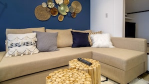 Ein Wohnzimmer mit einem beigefarbenen Sofa, mehreren Kissen, einem Holztisch und einem Wandbild.
