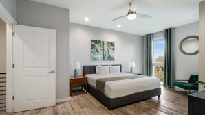 Une chambre moderne avec un grand lit, un ventilateur de plafond, une table de chevet, un miroir fixé au mur et une vue sur l’extérieur par une fenêtre.