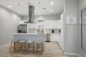 Une cuisine moderne avec des meubles blancs, un îlot central doté de tabourets, une hotte de cuisine en acier inoxydable et des luminaires suspendus.
