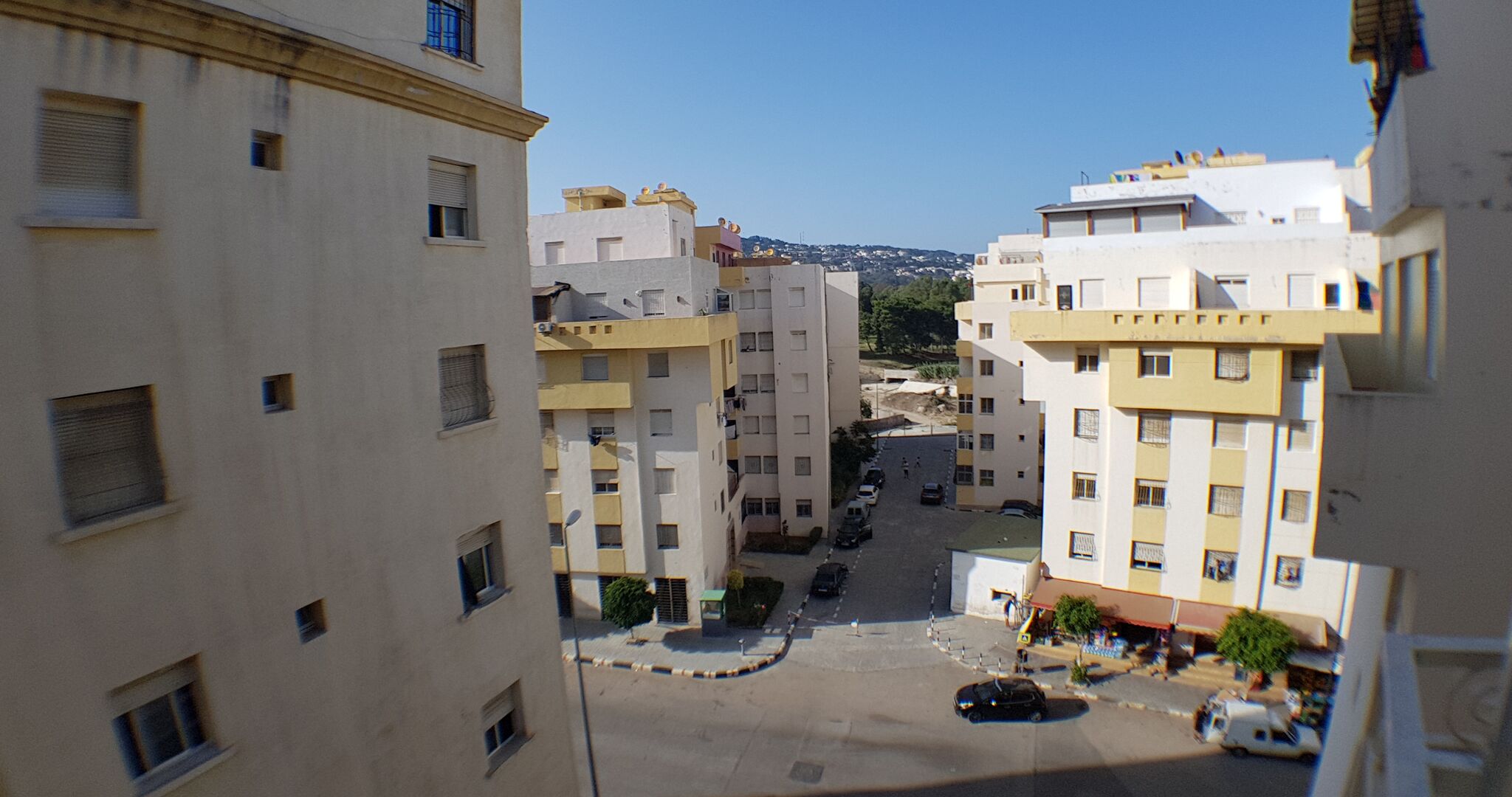 A narrow street between multi-story buildings with balconies, a clear sky, and distant hills.