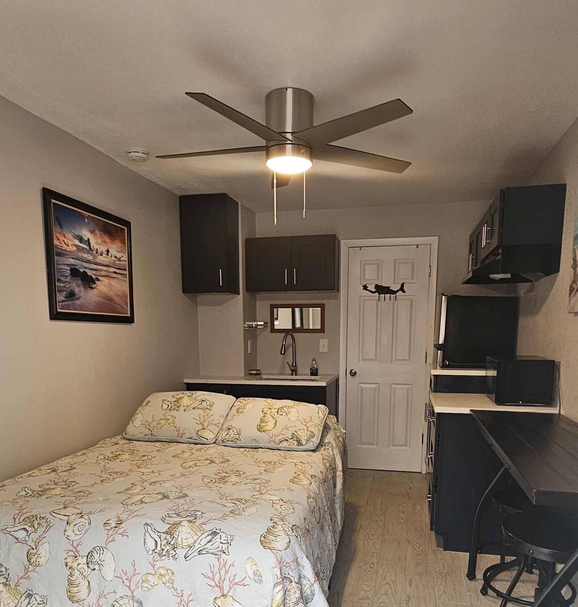 Une chambre compacte comprenant un lit, un bureau, une chaise, un ventilateur de plafond, une porte et une petite kitchenette.
