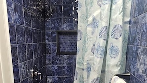 A shower area with a glass door, blue tiled walls, and a shower curtain with a seahorse and shell pattern.
