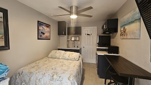 A compact room with a bed, a desk, a ceiling fan, and a door leading to a bathroom.