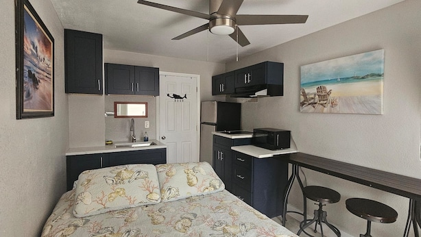 A compact bedroom with a bed, a small kitchenette, a ceiling fan, and a painting of a beach scene.