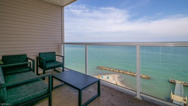 Un balcon avec deux chaises vertes et une table, donnant sur une plage protégée par une digue rocheuse.