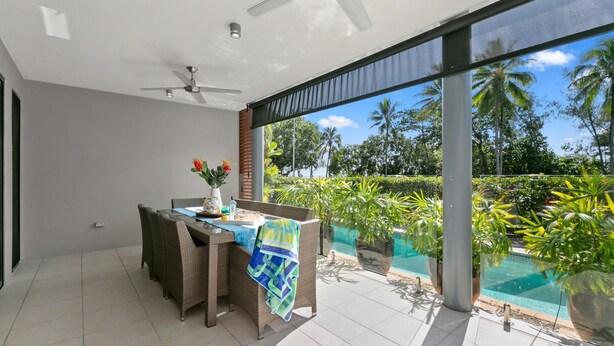 Un espace de restauration extérieur moderne, doté de mobilier en osier, d’un ventilateur de plafond et offrant une vue sur une piscine et une végétation tropicale.