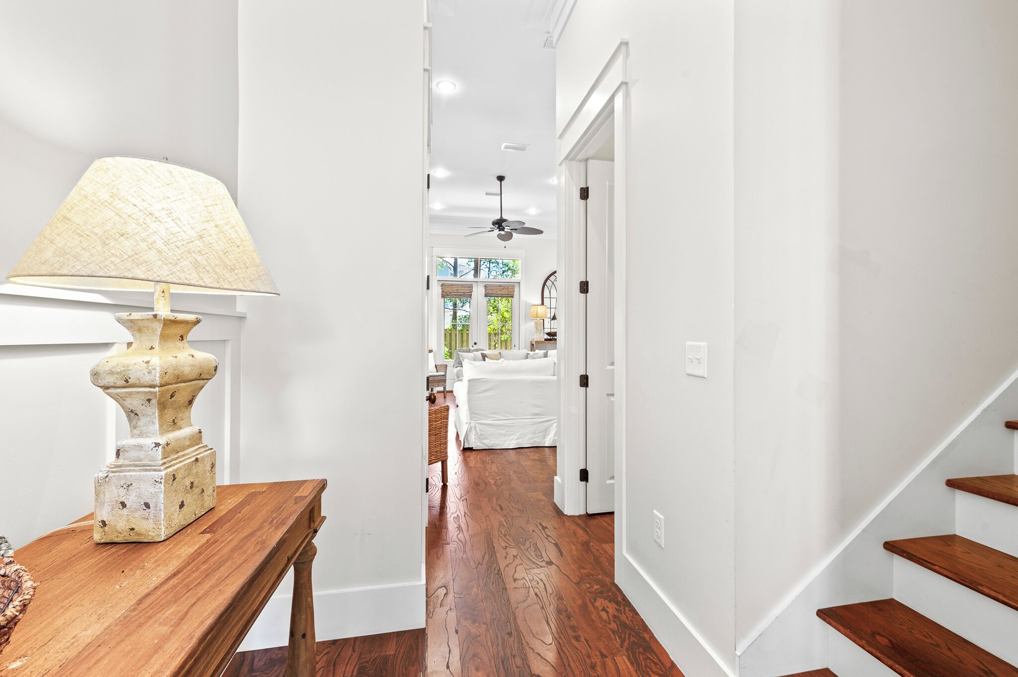 A hallway with a wooden table and lamp, leading to a bedroom with a bed and a ceiling fan.