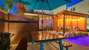An outdoor patio area with a wooden table and benches, a pergola with string lights, and a covered seating area with cushions. - 3-Level Brand New Townhouse in Mid City (New Orleans)