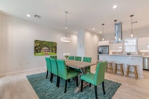 A modern dining area with a wooden table and green upholstered chairs, a kitchen with white cabinets, and a hanging light fixture.