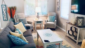 A living room with a grey sofa, two armchairs, a coffee table, a TV, and a window with blinds. - Charming Bungalow, remodeled, heated pool, walk to the beach (Naples)