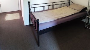 Ein Einzelbett mit Metallgestell und einer beigen Matratze in einem Zimmer mit weißen Wänden und Teppichboden.