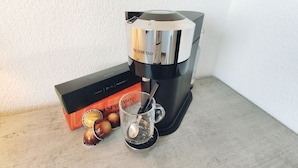 Eine Nespresso-Kaffeemaschine mit einer Packung, einer Glaskanne und einem Löffel auf einer Holzoberfläche.