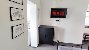 Ein Zimmer mit einem Bett, einem an der Wand montierten Fernseher, der 'NETFLIX' anzeigt, und gerahmten Kunstwerken an der Wand.