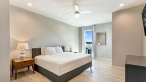 Une chambre d’hôtel moderne avec un grand lit, deux tables de chevet avec des lampes, un ventilateur de plafond et une vue sur la ville par une grande fenêtre.