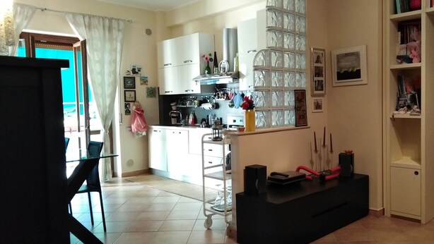 Een keuken met witte kasten, een zwart aanrecht en een glazen scheidingswand. Er is een eethoek met een tafel en stoelen, en een klein kastje met decoratieve spullen.