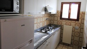 A compact kitchen with white cabinets, a built-in microwave, a gas stove, and a window with wooden frames. - villa MARISA 2 - Lignano (Lignano Sabbiadoro)