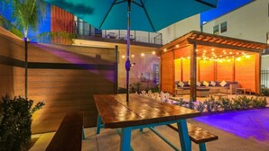 Un espace extérieur aménagé en terrasse, avec une table et des bancs en bois, une pergola illuminée de guirlandes lumineuses et un coin salon couvert agrémenté de coussins.