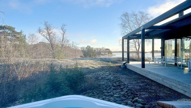 Un jacuzzi con vistas a una costa rocosa y árboles, una terraza de madera y un área de estar cubierta con bancos.