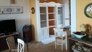 A living room with a television, a dining table with chairs, a bookshelf, and a decorative plant.