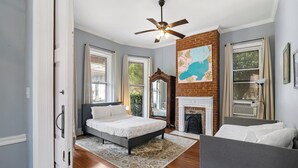 A bedroom with a bed, a fireplace, a ceiling fan, and a large window.