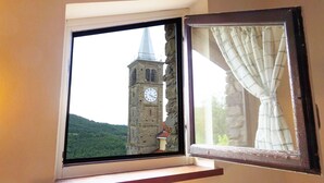 A window with a view of a stone tower with a clock, a curtain, and a small plant on a shelf. - Casa Clelia: the gem in the heart of the Langhe (Loazzolo)