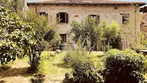 A stone building with a tiled roof, surrounded by a garden with various plants and a dirt path. - Casa Clelia: the gem in the heart of the Langhe (Loazzolo)
