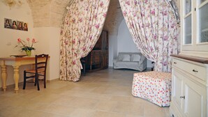 A room with a stone ceiling, floral curtains, a wooden chair, a bed with a floral bedspread, and a small table with a plant.