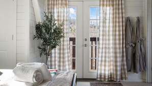 A room with a bed, a potted plant, checkered curtains, and a view through a door.