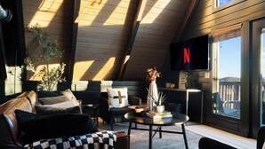A cozy living room with a high ceiling, a fireplace, a television, and a view of the outdoors.