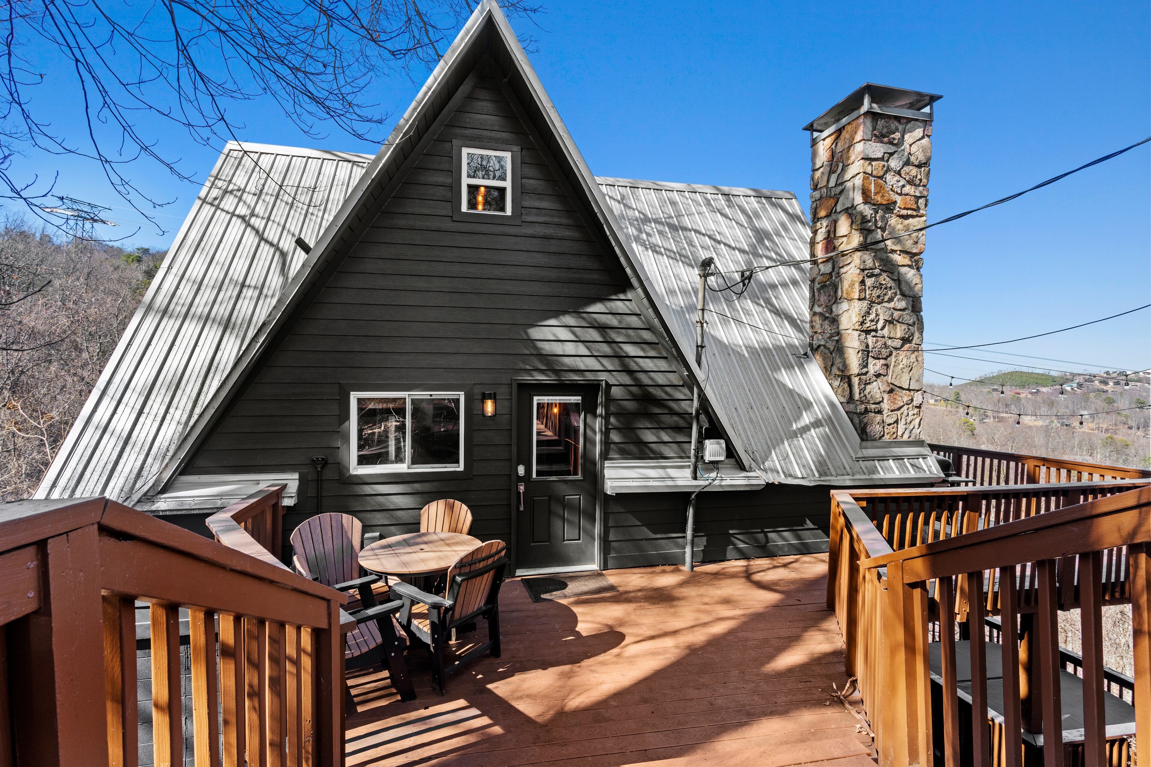 A dark wooden cabin with a stone chimney, a deck with chairs and a table, and a view of the surrounding landscape.