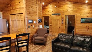 Un interior de cabaña acogedor con paredes de madera, una zona de comedor con mesa y sillas, un sofá de cuero y un televisor montado en la pared.