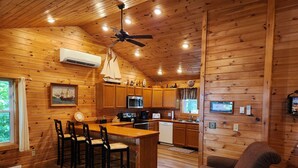 Interior de una cabaña con detalles de madera, una zona de cocina, un comedor con taburetes de bar, un sofá de cuero y un ventilador de techo.