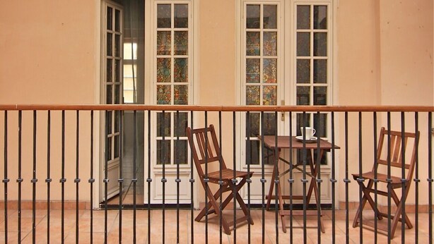 Uma varanda com guarda-corpo de madeira, duas janelas com vitrais e dois cadeiras de madeira com uma mesa pequena e uma xícara.