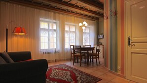 Uma sala de estar com sofá, mesa de jantar e cadeiras, tapete estampado vermelho e área de cozinha com máquina de lavar.