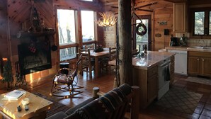 A cozy cabin interior with a fireplace, wooden dining table, rocking chair, and a kitchen area with a sink and countertop. - Pet-Friendly,Cozy Cabin,Hot Tub,Wood Fireplace,WiFi (Ferguson)