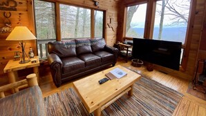 A cozy cabin interior with a leather sofa, wooden coffee table, and a television. Large windows offer views of the surrounding forest. - Pet-Friendly,Cozy Cabin,Hot Tub,Wood Fireplace,WiFi (Ferguson)