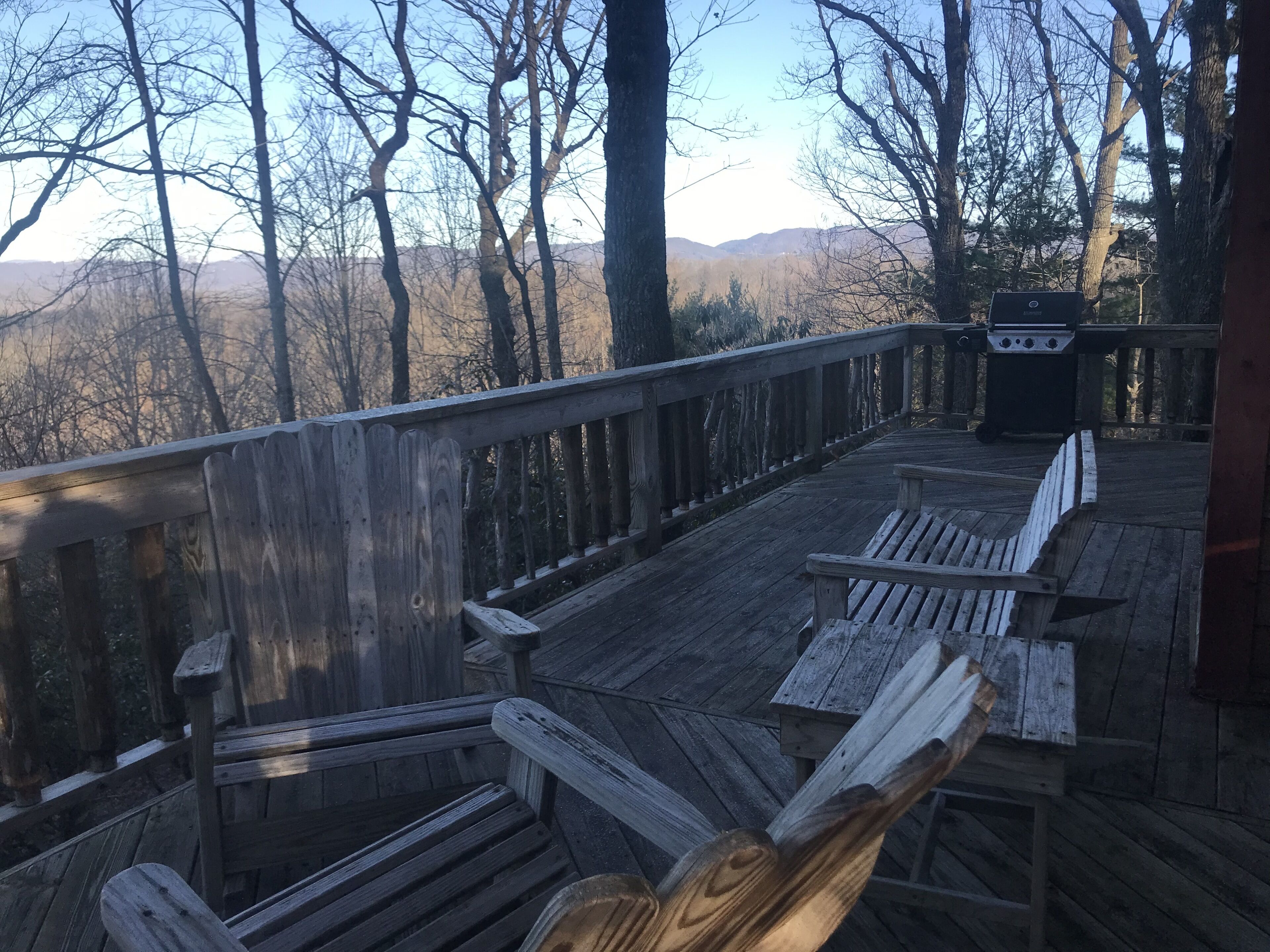 A wooden deck with benches and a grill, overlooking a forested area with mountains in the distance.