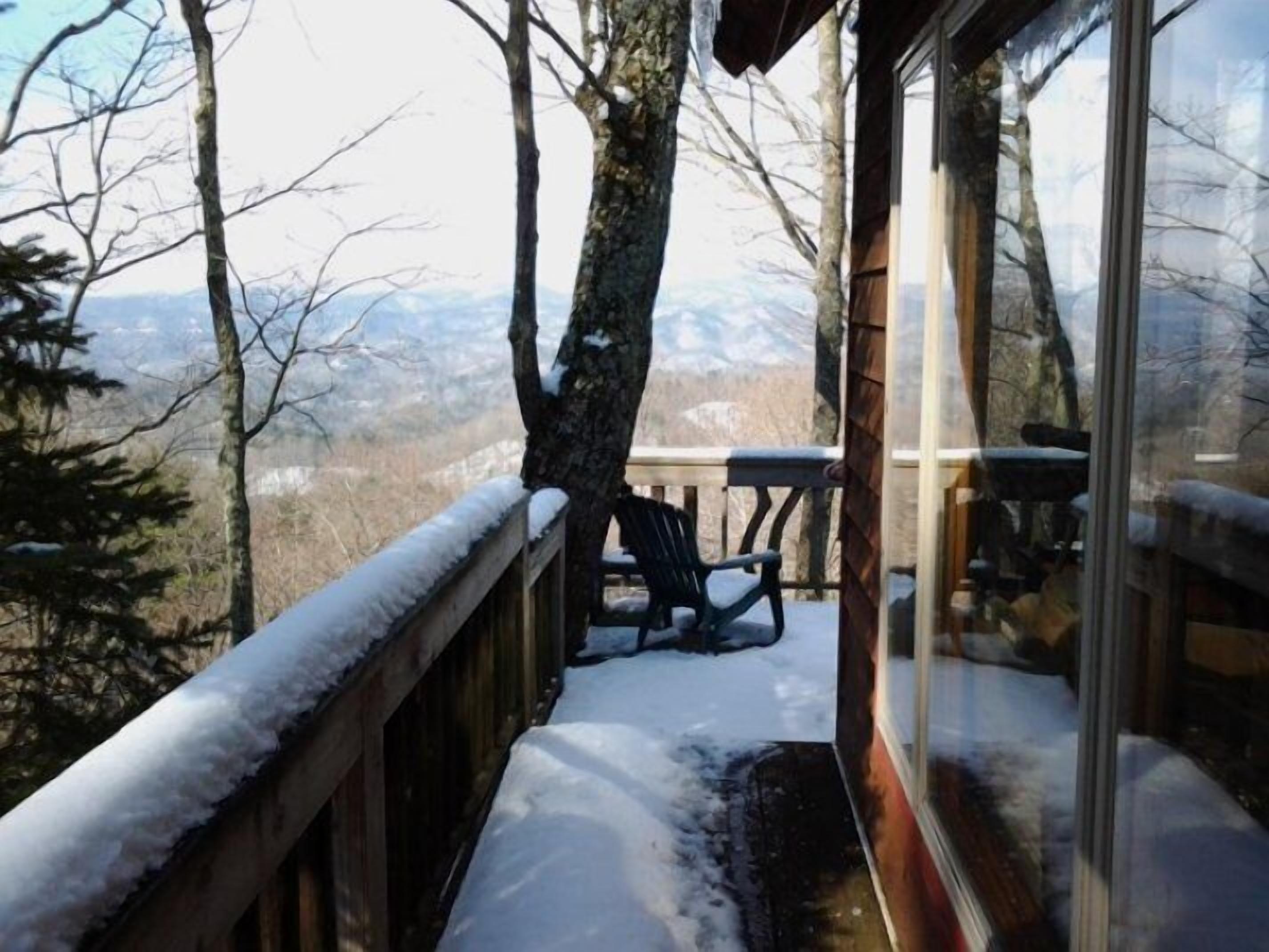 A wooden deck with snow-covered railings, a table, and chairs, overlooking a mountainous landscape.