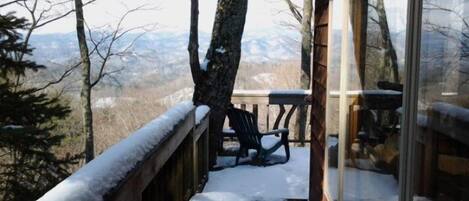 A wooden deck with snow-covered railings, a table, and chairs, overlooking a mountainous landscape.