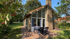 A small brick building with a glass door and windows, a brick pathway leading to a table and chairs, and a sign reading "DE BRANDING".