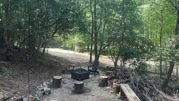 A forested campsite with a fire pit, benches, and a picnic table.