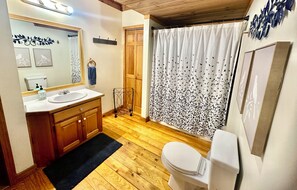 A bathroom with wooden flooring, a white toilet, a sink with a mirror, and a shower curtain with a leaf pattern. - Game Room, Fireplace and pit, very private, a little taste of heaven! (Dahlonega)