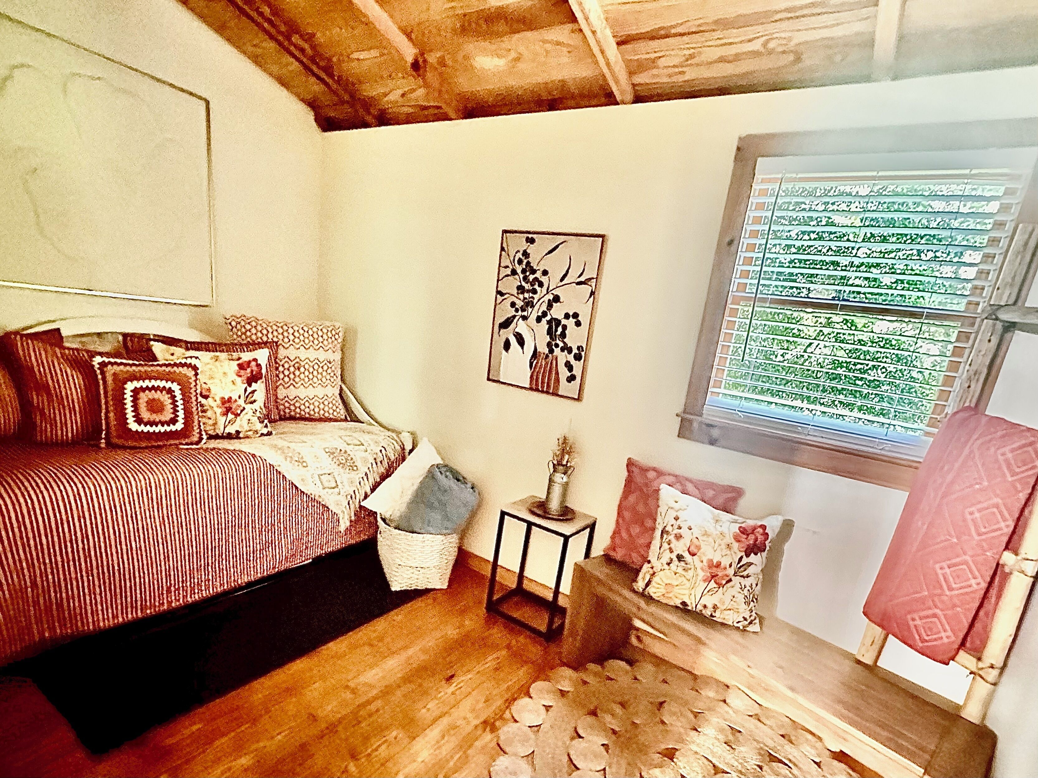 Een slaapkamer met een bed, kussens, een klein tafeltje, een aan de muur hangend schilderij en een raam met jaloezieën.