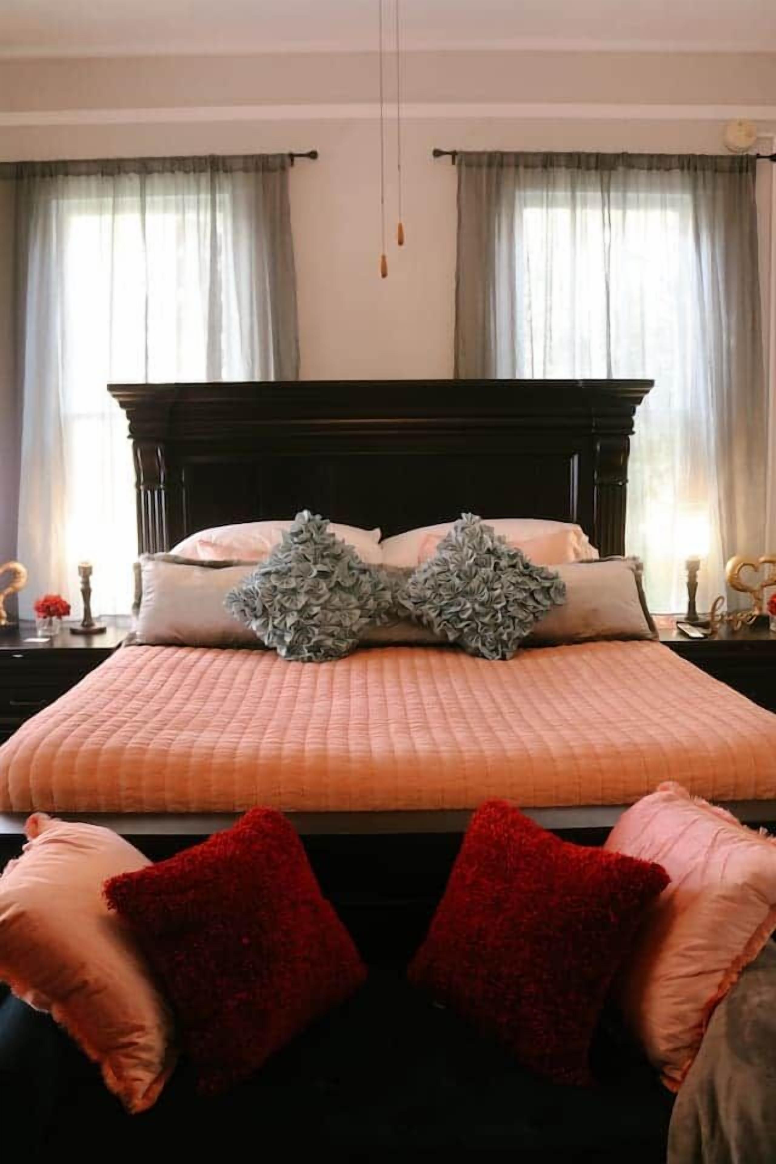A bedroom with a large bed, two windows with curtains, and a bedside table with a lamp.