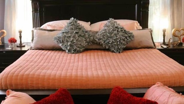 Un dormitorio con una cama grande, dos ventanas con cortinas y una mesita de noche con lámpara.
