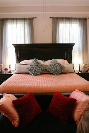 Un dormitorio con una cama grande, dos ventanas con cortinas y una mesita de noche con lámpara.