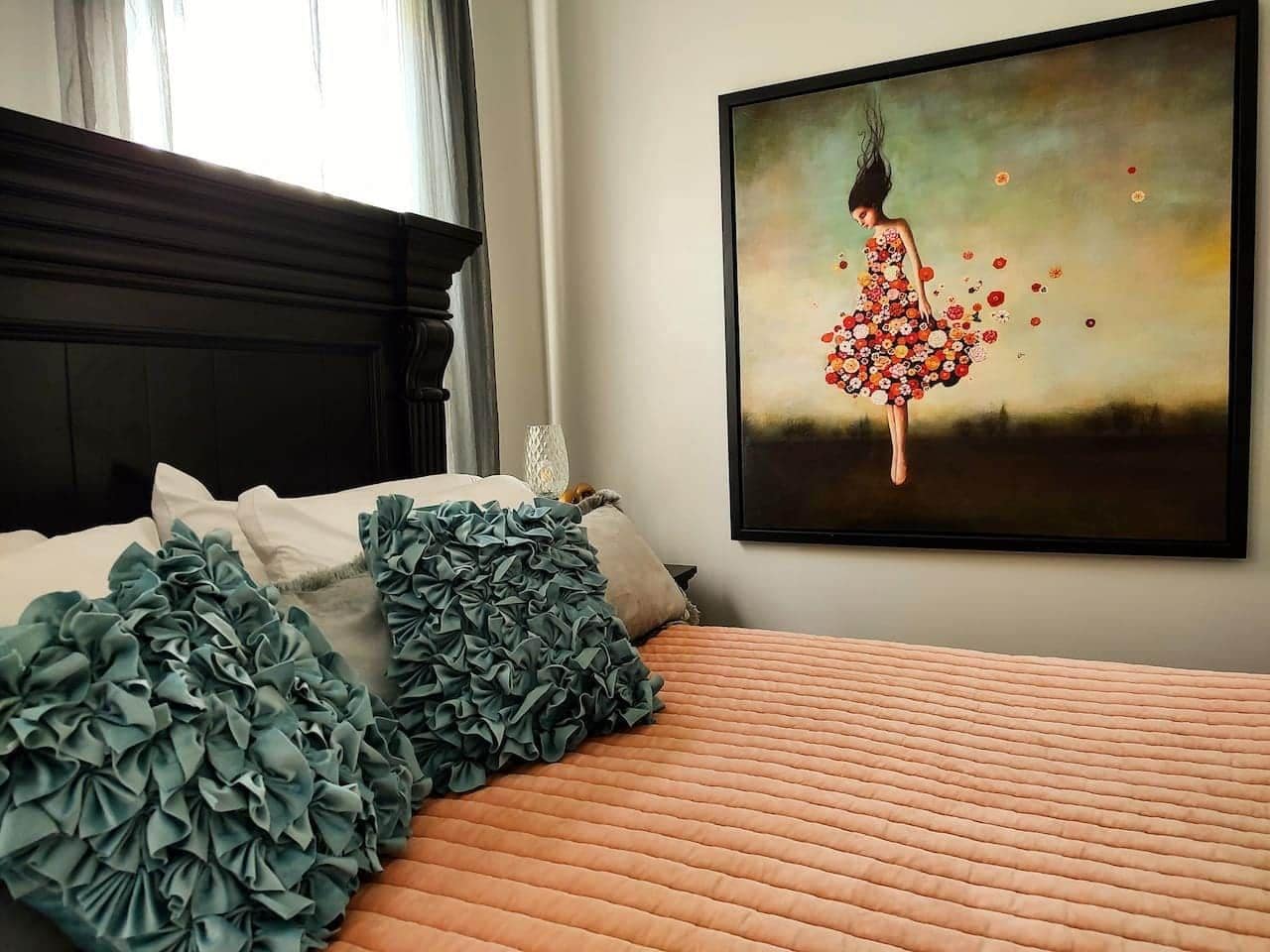 A neatly made bed with a patterned bedspread and decorative pillows, a painting of a woman in a floral dress on the wall, and a window with curtains.