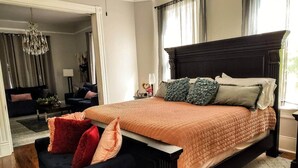 Un dormitorio con una cama grande, un cabecero oscuro y una pequeña zona de estar con una silla y un reposapiés.
