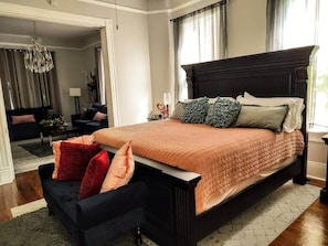 Un dormitorio con una cama grande, un cabecero oscuro y una pequeña zona de estar con una silla y un reposapiés.