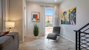 A modern living room with a grey sofa, a floor lamp, a potted plant, and a black chair. There is a painting on the wall and a window with a view of buildings.